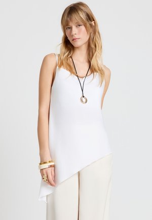 Femme blonde portant un top blanc asymétrique sans manches, pantalon crème, collier en plusieurs couches, bracelets et bague en or, debout devant un fond gris.