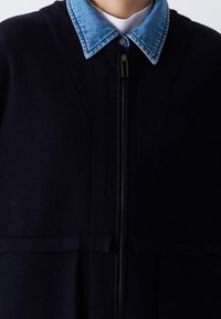 Tmavě modrý kabát z vlněné směsi s denimovým límcem, plným zapínáním na zip a dvěma horizontálními švy v pase. Texturovaná tkanina s hladkým povrchem.