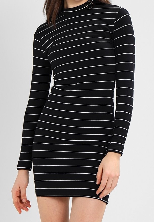 Femme portant une robe noire cintrée à manches longues avec de fines rayures blanches horizontales, debout devant un fond uni.