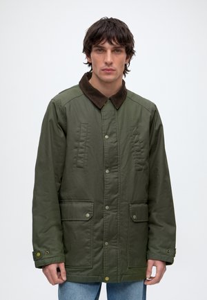 Homme portant une veste verte boutonnée avec un col marron et des poches avant à rabat, debout sur un fond blanc uni.