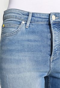 Vaqueros de mezclilla de un azul claro con un ajuste Slim. Cuenta con cinco bolsillos, costuras amarillas y un cierre de botón plateado. Textura suave y ligeramente desgastada.