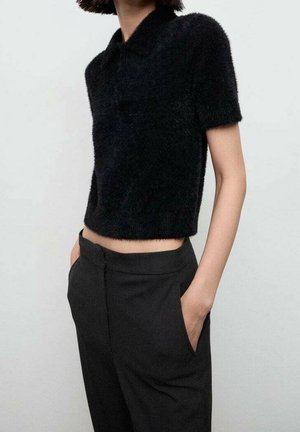 Frau trägt ein kurzärmeliges, flauschiges schwarzes Croptop und hoch taillierte schwarze Hosen, die Hände in den Taschen, vor einem schlichten Hintergrund.