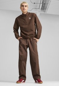 Brun zip-up jacka och matchande byxor, båda med en texturerad tyg, med en Puma-logotyp. Modellen bär röda och vita sneakers.