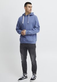 Felpa blu con cappuccio e lacci bianchi, abbinata a jeans grigio scuro e sneakers nere. Look casual e comodo con polsini a coste.