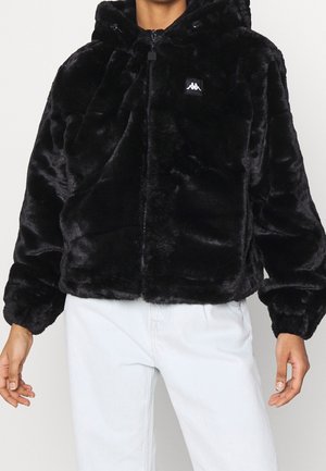 Person iført en sort, blød hættejakke med lynlås foran og hvidt logo påsyet, kombineret med lyseblå jeans.