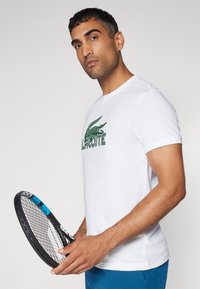 Camiseta de algodón blanca con un logo de cocodrilo verde y texto. La persona sostiene una raqueta de tenis con acentos en negro y azul.
