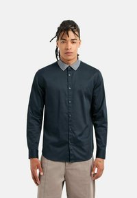 Camicia blu navy con colletto grigio chiaro a contrasto. Maniche lunghe, texture liscia e logo discreto sul petto.