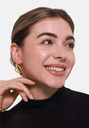 Orecchini d'oro presentano un design intrecciato con una superficie liscia. Il modello li indossa con un dolcevita nero.