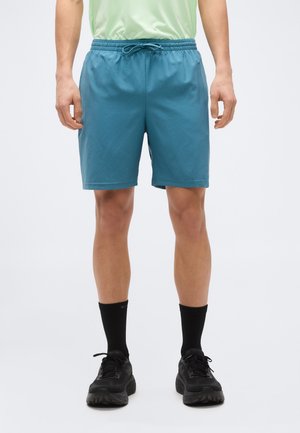 Homme portant un short de sport bleu sarcelle avec cordon de serrage, des chaussettes noires montantes et des chaussures de course noires, debout devant un fond blanc uni.
