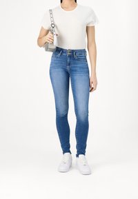 Persona vestida con una camiseta blanca, jeans skinny de tiro alto azules con doble botón, zapatillas blancas y sosteniendo un pequeño bolso plateado de hombro.