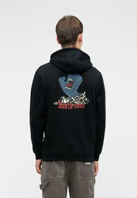 Crna dukserica s šarenim grafičkim prikazom plave ruke s ustima, preko pozadine stiliziranih oblika, sa tekstom "SANTA CRUZ" ispod.