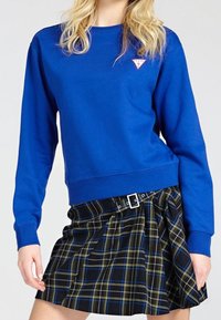 Mujer que lleva un sudadera azul de mangas largas con un pequeño logo y una falda de cuadros negra con pliegues, con rayas azules y amarillas y una hebilla plateada.