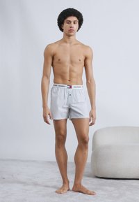 Λευκά βαμβακερά μποξεράκια με ελαστική μέση που φέρει το λογότυπο «TOMMY HILFIGER». Το μοντέλο στέκεται μπροστά σε μονόχρωμο φόντο, χωρίς να φορά πουκάμισο.