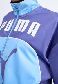 Man som bär en lila och ljusblå Puma zip-up jacka med vit Puma-logotyp och text över bröstet och kragen.