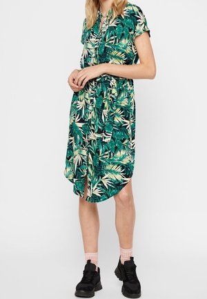 Vrouw draagt een groene jurk met tropische bladprint, gecombineerd met zwarte chunky sneakers en lichtroze sokken, staand tegen een effen achtergrond.