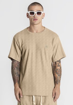 Hombre con camisa de manga corta beige con estampado y pantalones cortos a juego, gafas de sol blancas, con tatuajes que cubren ambos antebrazos.