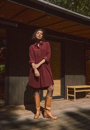 Femme en robe bordeaux et bottes hautes beige debout sur une terrasse en bois devant un bâtiment en bois foncé sous le soleil.