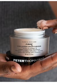 Tarro plateado etiquetado como "FIRMx Collagen Moisturizer" con base negra. Crema espesa en la yema del dedo. Textura suave, tamaño 50 ml, marca de cuidado de la piel clínica.
