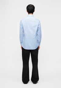 Άνδρας που στέκεται γυρισμένος πίσω, φορώντας ένα ανοιχτό μπλε πουκάμισο με μακριά μανίκια και μαύρο παντελόνι με φαρδιές παντελόνες, μπροστά σε απλό λευκό φόντο.