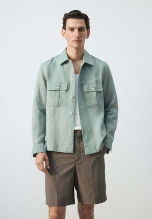 Hombre con camisa verde claro abotonada sobre camiseta blanca y pantalones cortos marrones de rayas delgadas, de pie frente a un fondo liso.