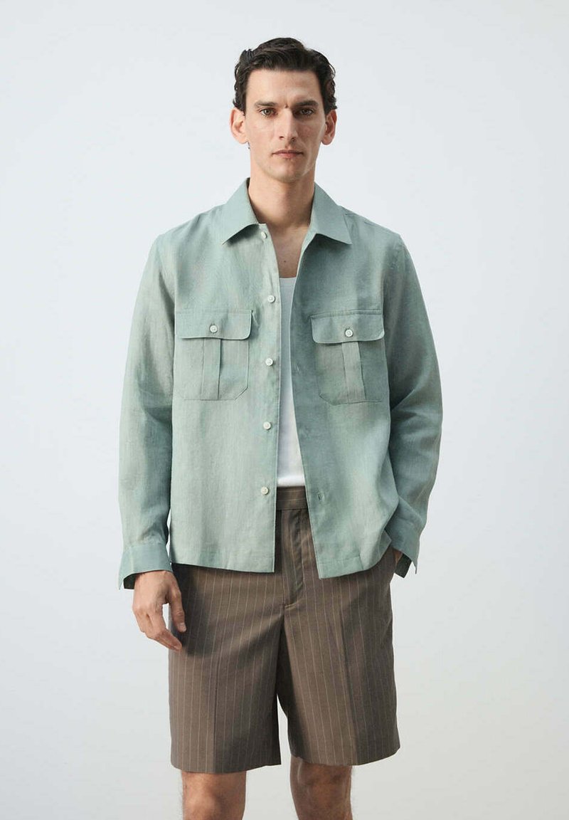 Homme portant une chemise vert clair boutonnée sur un haut blanc et un short marron à fines rayures, debout devant un fond uni.