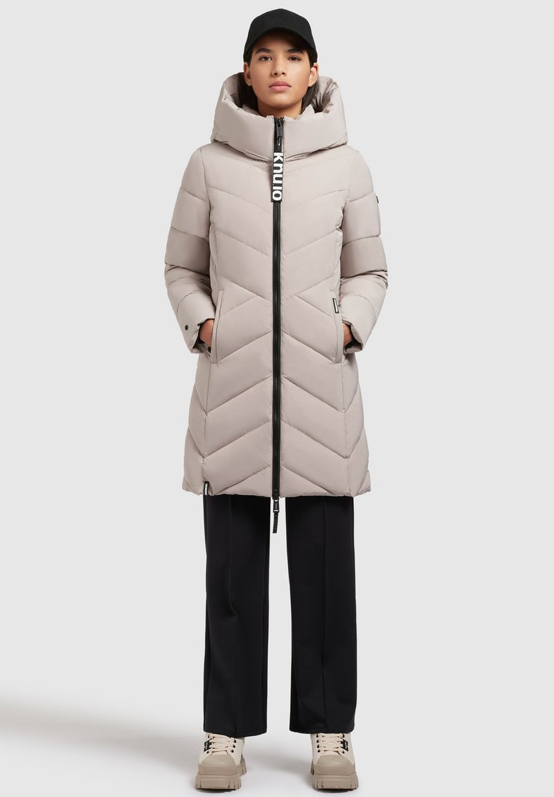 Ljust beige puffjacka med ett chevronmönster, hög krage, dragkedjeförslutning och sidofickor. Kombineras med mörka byxor och beiga sneakers.