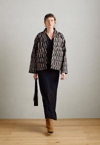 Veste matelassée à motifs noir et blanc sur une robe noire, assortie de bottines à talons beiges et d'un sac noir, sur un parquet en bois clair.