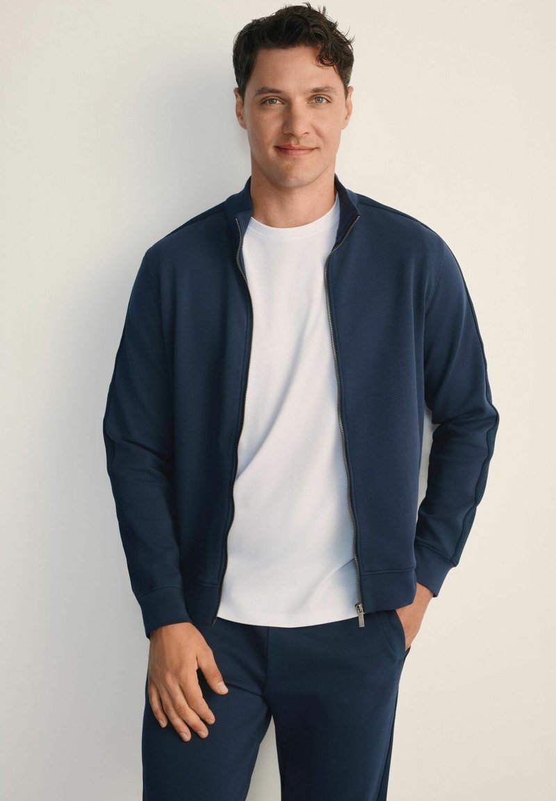 Granatowa kurtka na zamek z gładką teksturą i stojącym kołnierzem, noszona na białym t-shircie z okrągłym dekoltem. Gładki materiał, minimalistyczny design.