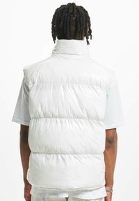 White puffy vest with a high collar, featuring quilted sections and a shiny texture. The design is sleeveless, with a relaxed fit.