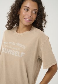 T-shirt en coton beige à manches courtes, col rond et texte en strass indiquant "LA VRAIE CROISSANCE EST D'ÊTRE DAVANTAGE NOUS-MÊMES."