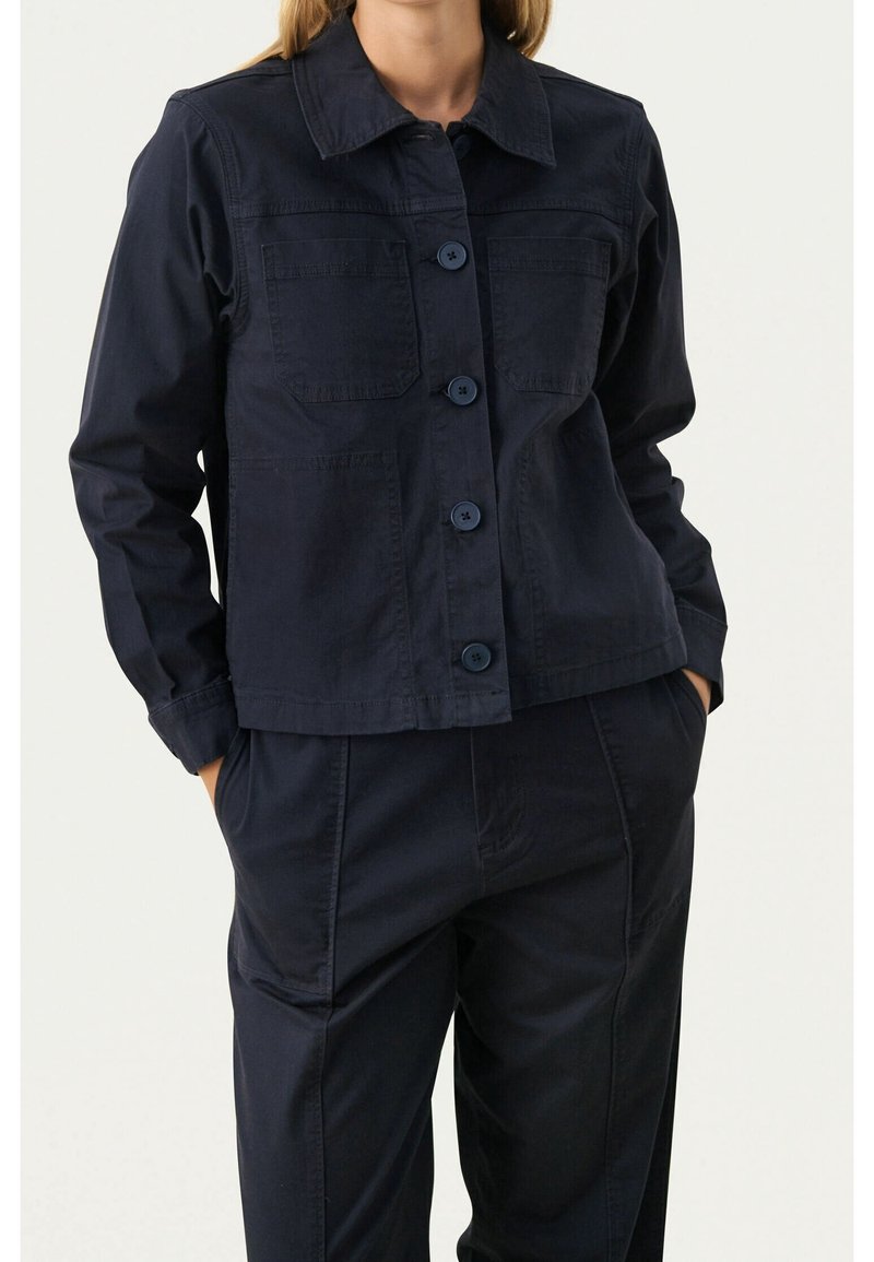 Femme portant une veste boutonnée bleu marine foncé avec poches sur la poitrine et pantalon assorti, mains posées de manière décontractée dans les poches du pantalon, fond neutre.