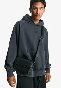 Svart axelremsväska med lock, texturerad yta och justerbar rem, ihop med en kolgrå hoodie med känguruficka och logotyp.