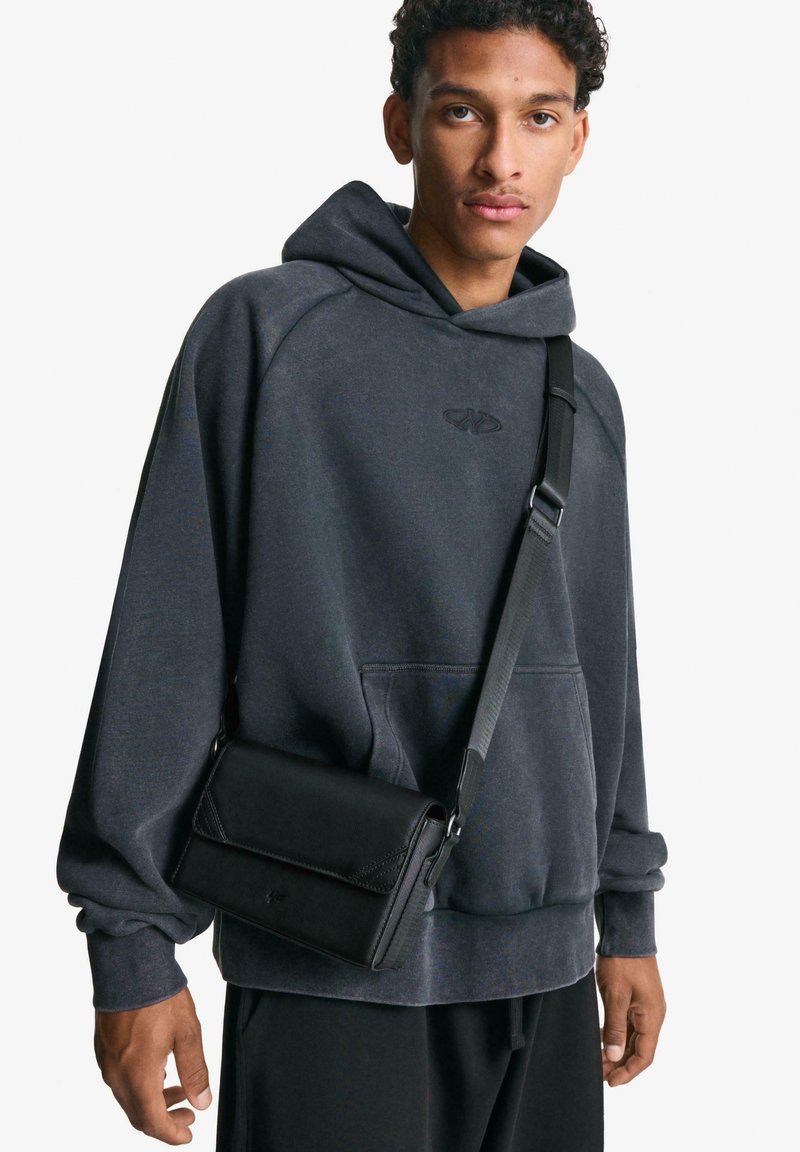 Svart axelremsväska med lock, texturerad yta och justerbar rem, ihop med en kolgrå hoodie med känguruficka och logotyp.