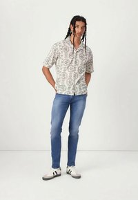 Ung man med långt flätat hår som bär en kortärmad mönstrad skjorta, blå slim-fit jeans och vita sneakers stående mot en vit bakgrund.