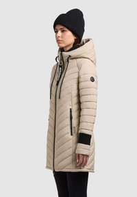 Beige isolierter Mantel mit Kapuze, diagonal gesteppt, zwei Reißverschlusstaschen und einem gerippten schwarzen Bündchen. Schwarze Mütze auf dem Kopf des Modells.