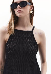 Top nero senza maniche con un motivo a zigzag testurizzato, realizzato in materiale a maglia. Gli accessori includono occhiali da sole ovali neri e orecchini a forma di stella dorati.