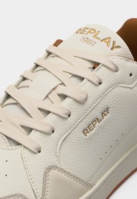 Gros plan sur une chaussure de sport en cuir blanc avec des lacets beiges, la marque « REPLAY 1981 » sur la languette et « REPLAY » embossé sur le panneau latéral.