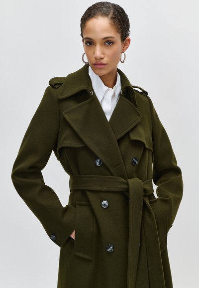 Casaco verde-oliva de dois botões com cintura cingida, gola larga e bolsos laterais; feito de um tecido texturizado, apresentando grandes botões pretos.