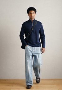 Tengerészkék polár cipzáros pulóver, világoskék bőszárú farmernadrág, barna mokaszinok és fekete oldaltáska, semleges háttér előtt.