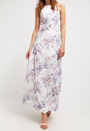 Kobieta ubrana w białą, bezrękawnikową sukienkę do ziemi z pastelowym, kwiatowym akwarelowym wzorem i beżowe, odkryte buty na obcasie.