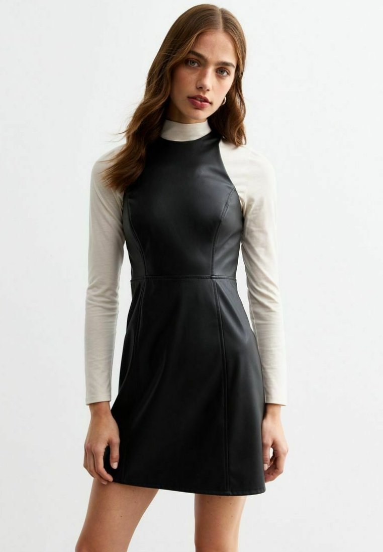 Abito in ecopelle nera con scollatura alta e design senza maniche, sovrapposto a una maglietta a maniche lunghe color crema. Texture liscia e forma strutturata.