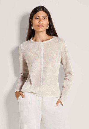 Vrouw met lang donker haar draagt een lichtbeige, geknoopte cardigan met bloemmotief en witte broek, handen in de zakken, neutrale achtergrond.