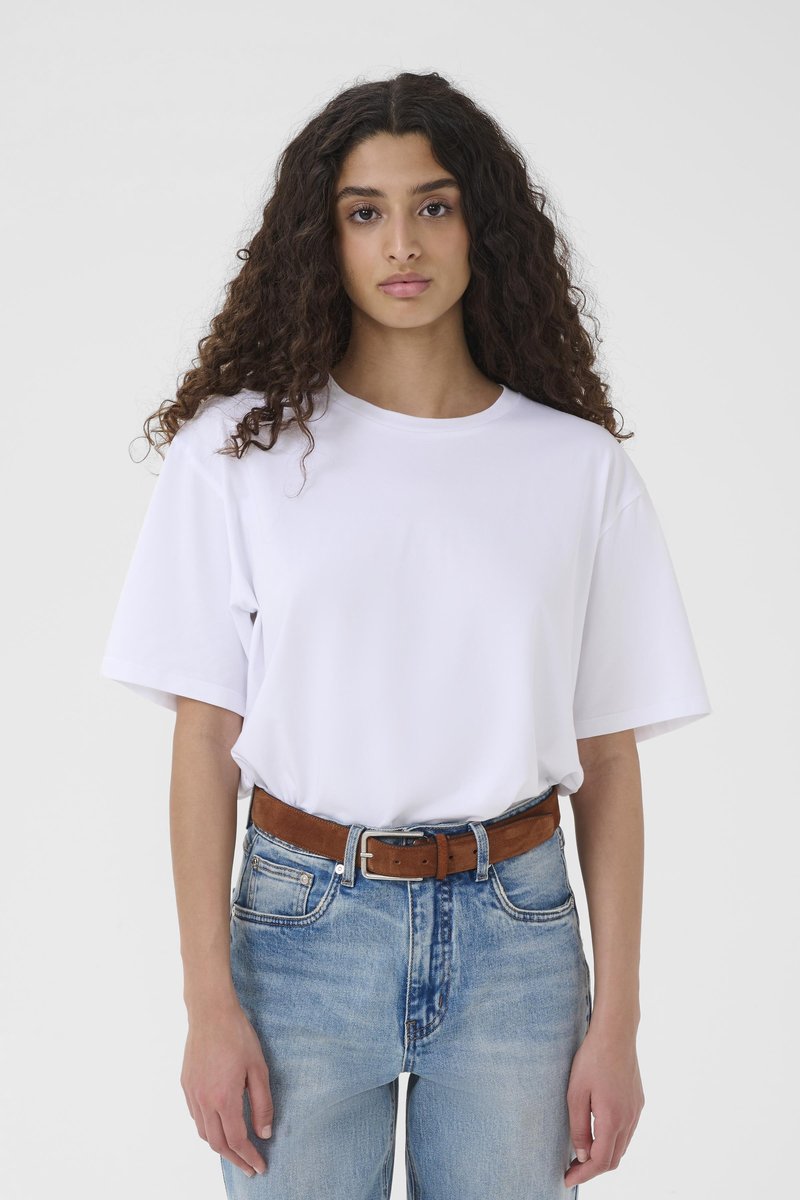 Young woman with long curly hair wearing a plain white oversized T-shirt tucked into blue jeans with a brown belt, standing against a white background.