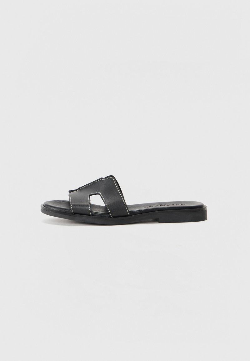 Black leather slide sandal with a flat sole, featuring two crossed straps and contrasting white stitching. Simple open-toe design.
