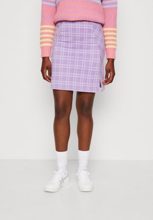 Personne portant une jupe à carreaux violette avec une fente latérale, des baskets blanches, des chaussettes blanches et un pull rayé rose, debout devant un fond blanc.