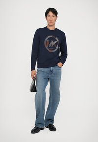 Sweatshirt azul-marinho com um design de logótipo circular em laranja e cinza, combinado com jeans claros de perna larga e sapatos pretos; segurando uma mala preta.