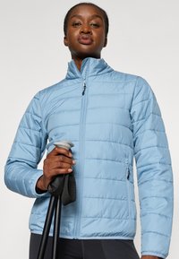 Giacca isolante azzurro chiaro con chiusura a zip, cuciture orizzontali, tasche laterali e colletto alto. Modello che tiene bastoncini da trekking.