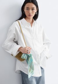 Jeune femme portant une chemise blanche surdimensionnée et un jean, tenant un sac à bandoulière marron orné d'un foulard bleu à motifs.