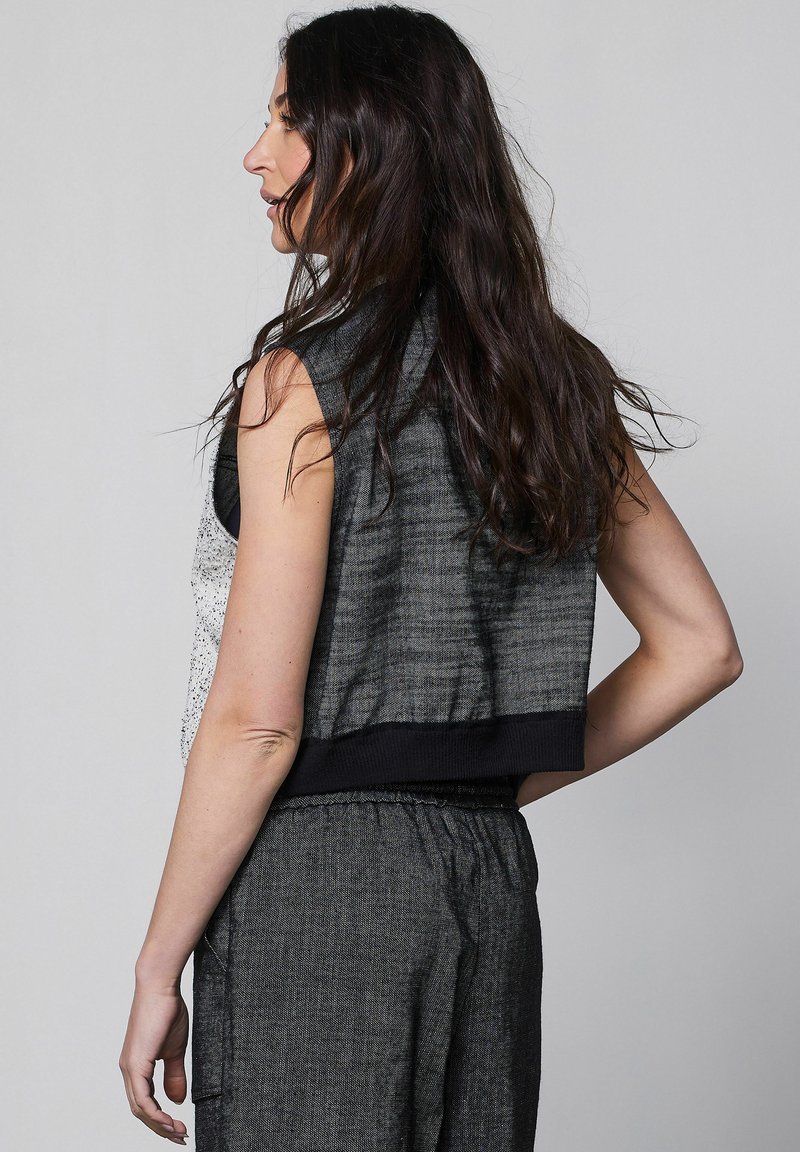 Femme aux longs cheveux foncés portant un haut sans manches gris foncé et un pantalon texturé assorti, debout de profil sur un fond uni.