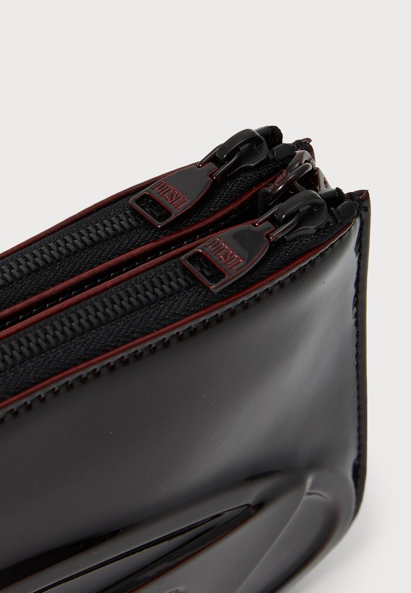 Pochette noire brillante avec des fermetures à double zip et des détails en tissu texturé. Présente des accents rouges contrastants et des contours lisses.